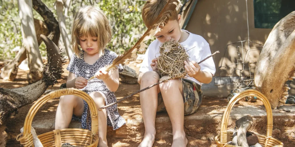 Duas crianças segurando capim seco durante atividade educativa em acampamento de safári com rochas e natureza ao redor
