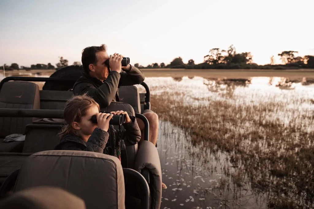 Pai e filha em veículo de safári aberto usando binoculos para observar vida selvagem, com paisagem de savana alagada ao fundo