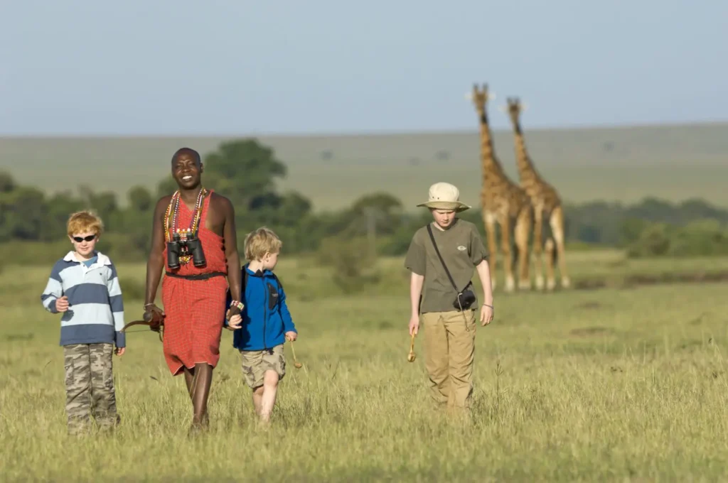 Guia em traje tradicional vermelho caminhando com crianças em savana verde, com girafas ao fundo durante caminhada na natureza