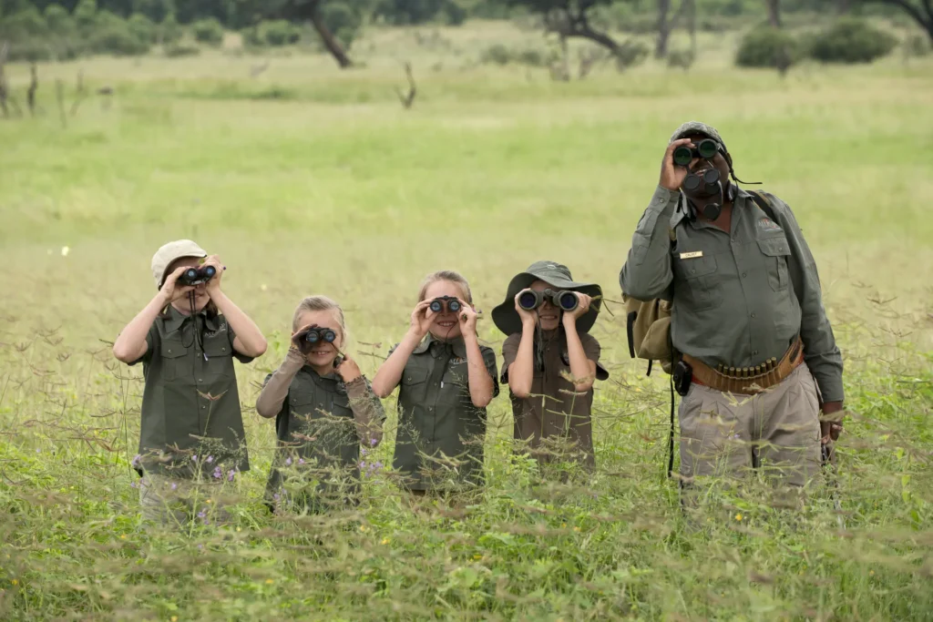 Guia de safári com crianças e adultos alinhados usando binoculos para observar e identificar vida selvagem em savana verde