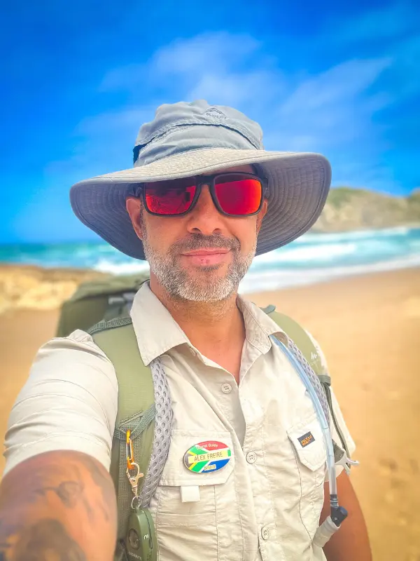 Retrato de um guia de safari profissional com chapéu de abas largas, óculos vermelhos e insígnia da África do Sul, com praia ao fundo.