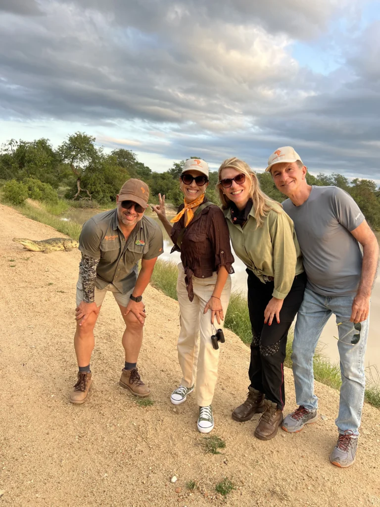 Guia de safári com grupo de quatro turistas posando para foto em caminho de terra, com savana verde e céu nublado ao fundo