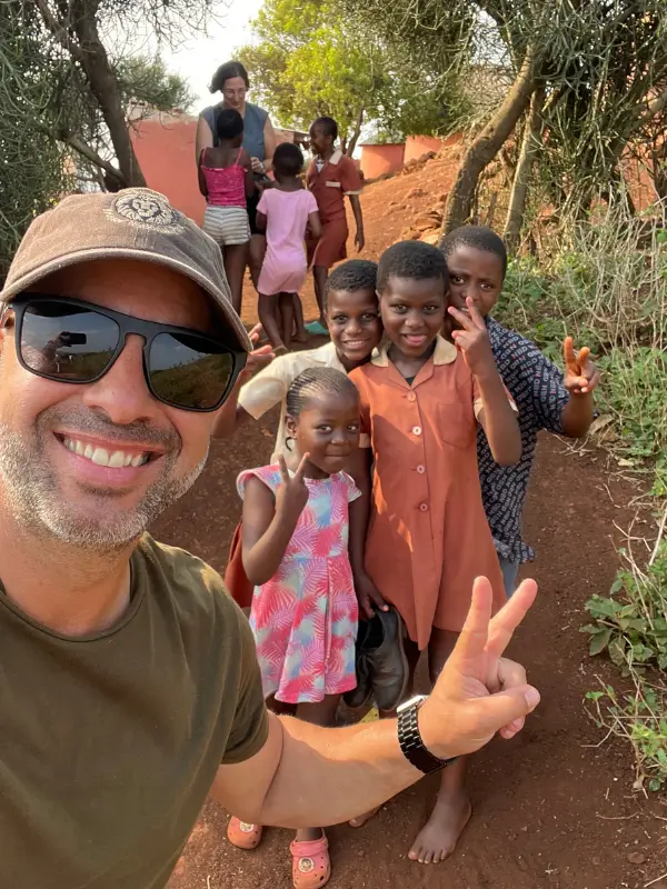 Selfie de um homem em uniforme de safari com grupo de crianças locais sorridentes em uma aldeia, todos acenando para a câmara.