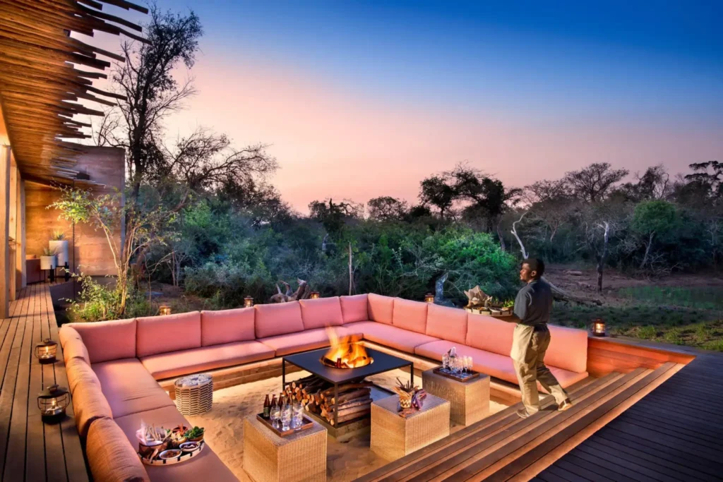 Área de estar de lodge de luxo ao entardecer com sofá em forma de L em tom coral, fogueira central acesa e vista panorâmica para savana verde ao fundo