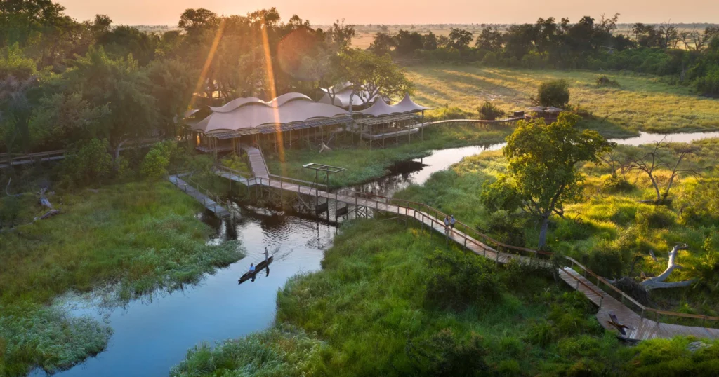 Vista aérea de lodge de safári com tendas e estruturas ao entardecer, localizado junto a rio sinuoso rodeado por savana verde e arvoredo denso