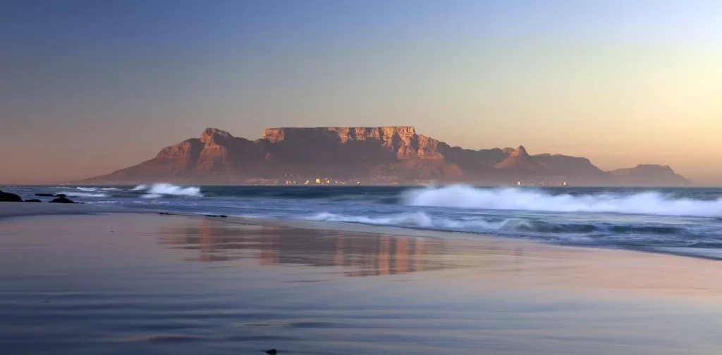 Tabela Montanha ao nascer do sol refletida na praia molhada com ondas quebrando na areia branca.