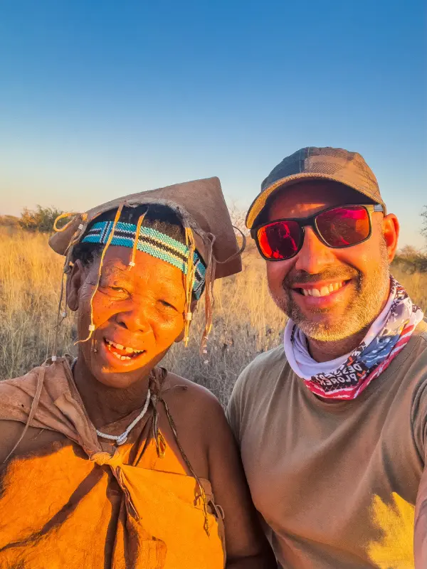 Selfie de um homem e uma mulher guia local com pintura facial laranja e fita de contas tradicional, ambos sorrindo contra fundo de savana seca.