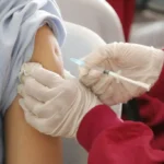 Profissional de saúde com luvas brancas administrando injeção de vacina no braço de pessoa envolvida em toalha de proteção branca