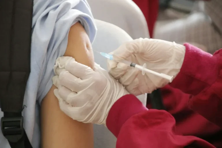 Profissional de saúde com luvas brancas administrando injeção de vacina no braço de pessoa envolvida em toalha de proteção branca