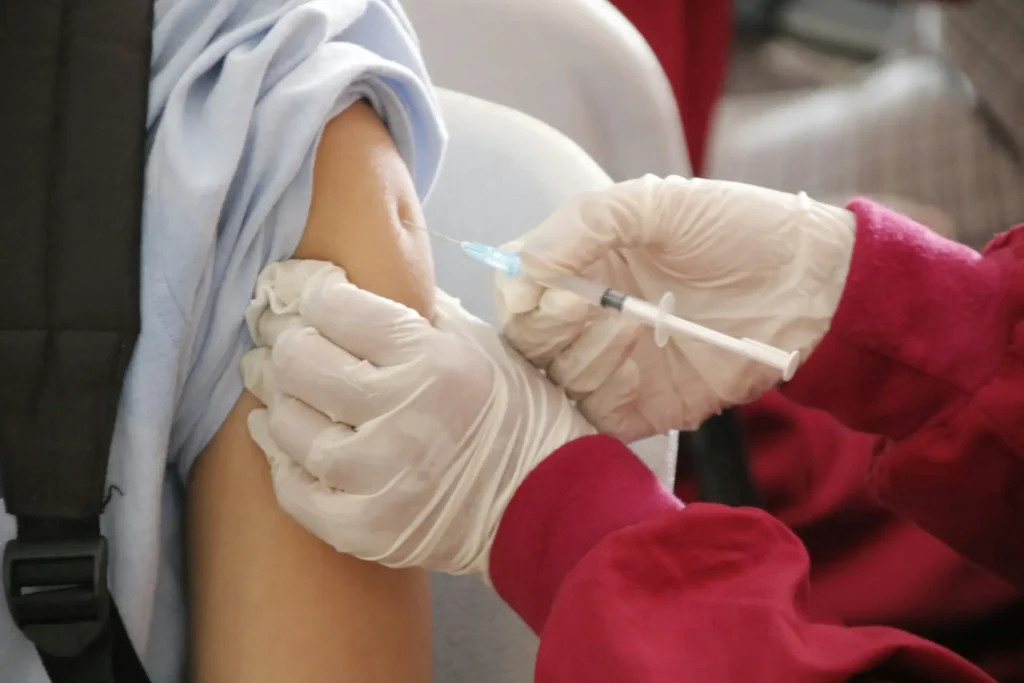 Profissional de saúde com luvas brancas administrando injeção de vacina no braço de pessoa envolvida em toalha de proteção branca